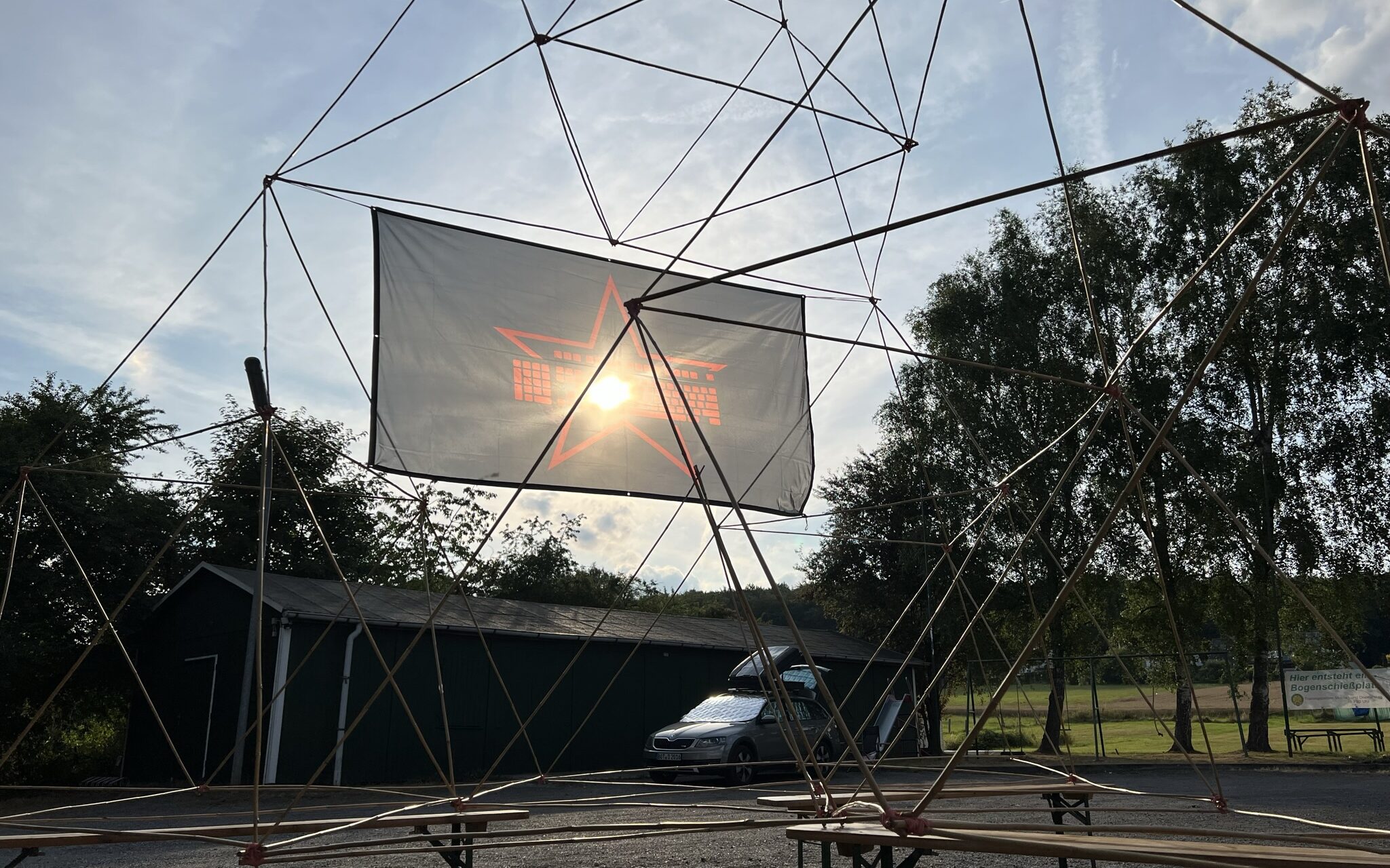 Aus Bambusstangen und Verbindern gebaute Struktur im Sonnen-Gegenlicht. Das Sonnenlicht wird von einer Tastatur-Stern-Flagge etwas gedämpft.