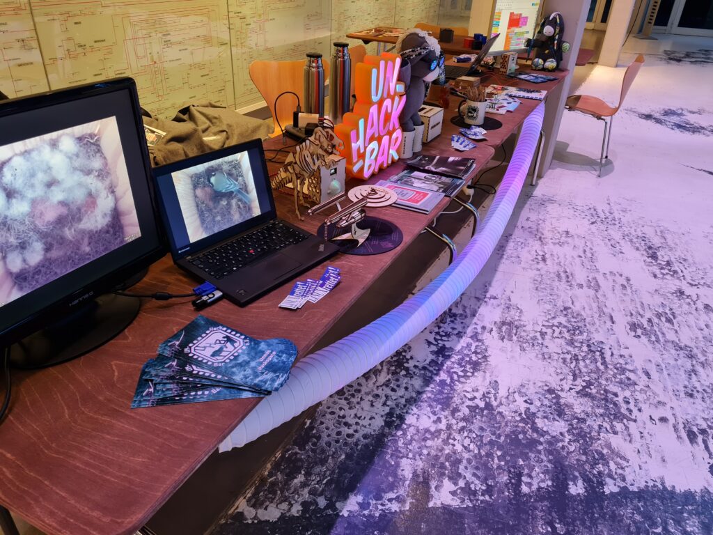 Stand der UN-Hack-Bar auf der Maker Faire Ruhr 2022.
Man sieht: 2 Monitore mit Bilder aus einem Nistkasten, Flyer, Aufkleber, CO2 Ampel, ein leuchtender Schriftzug "UN-Hack-Bar", ein Stofftier-Esel, UNHB Tassen, Datenschleuder-Zeitschriften 