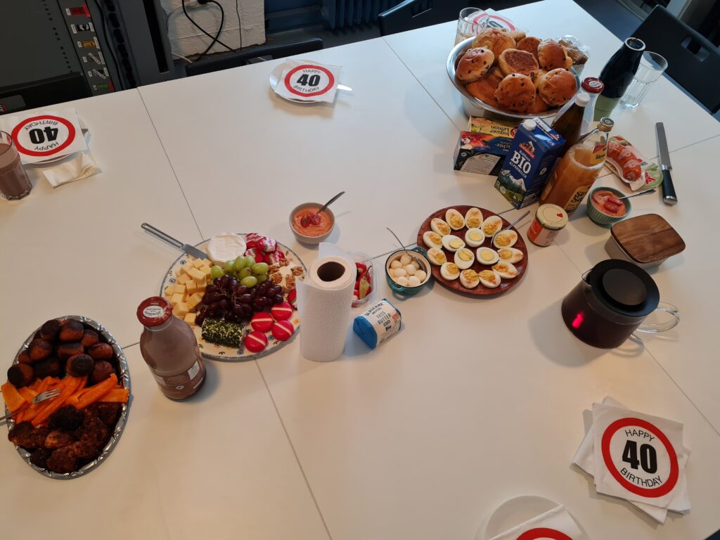 Ein Tisch der div. Frühstücks-Nahrungsmitteln gedeckt ist. So finden sich zB. Eier, eine Käseplatte, Gemüse, Tee, Brötchen, Milch und Säfte. Außerdem Servitten mit "Happy 40 Birthday" im Stil eines "Höchstgeschwindigkeit"-Verkehrsschild.