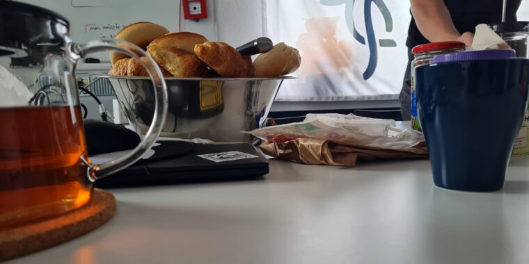 Foto aus der Froschpersektive von einem gedeckten Frühstückstisch. Zu sehen sind Brtöchen, eine Kanne Tee, eine Tasse, Aufschnitt aber auch ein Laptop und ein Whitboard mit etwas schwer zu lesenden Text und eine Flagge mit Chaosknoten