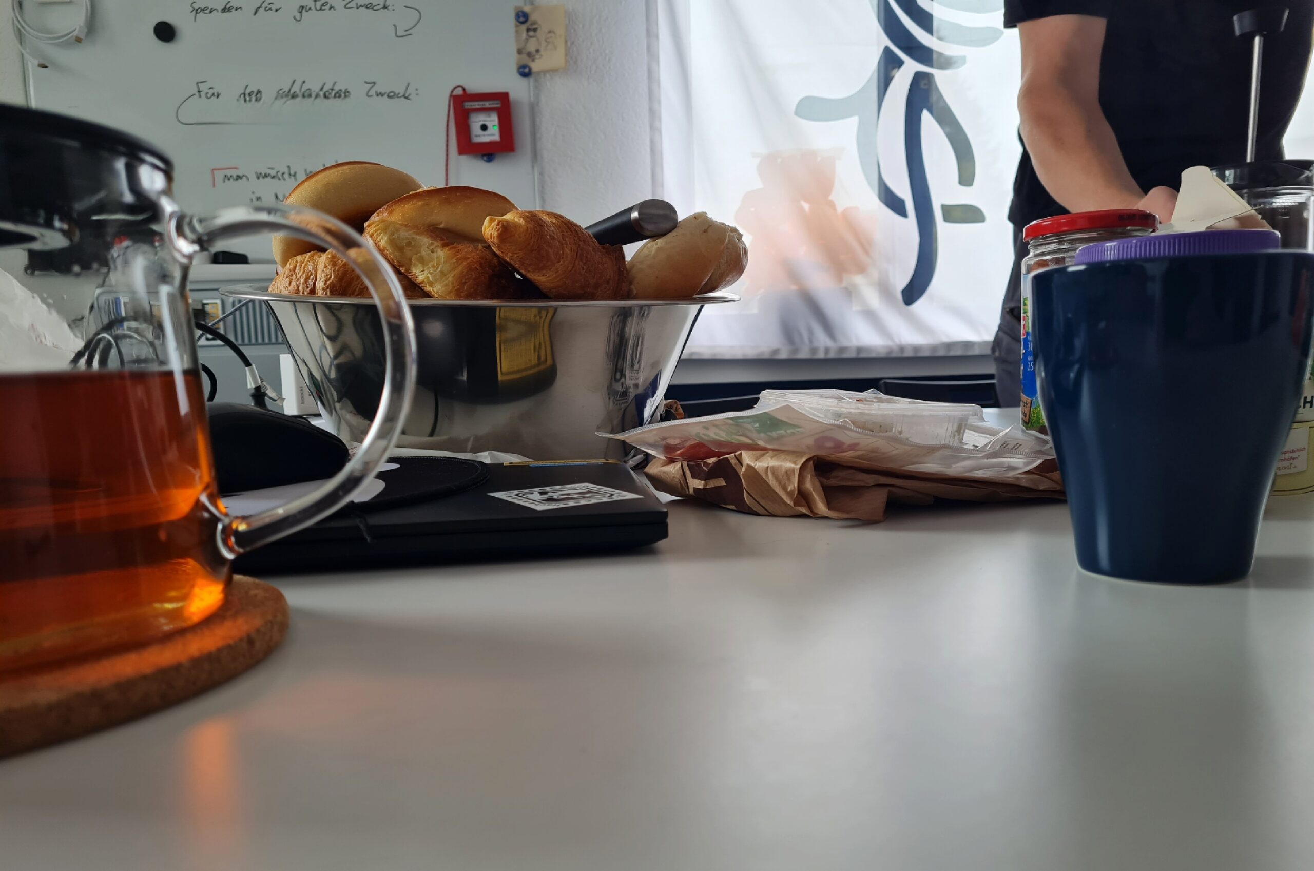 Foto aus der Froschpersektive von einem gedeckten Frühstückstisch. Zu sehen sind Brtöchen, eine Kanne Tee, eine Tasse, Aufschnitt aber auch ein Laptop und ein Whitboard mit etwas schwer zu lesenden Text und eine Flagge mit Chaosknoten