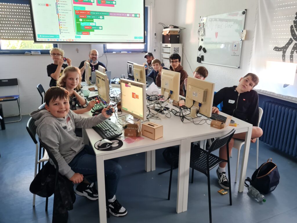 Foto von 7 Kindern und Jugendlichen die an PCs sitzen und jeweils einen Calliope Mini programmieren. Im Hintergrund noch der Dozent und auf dem Beamer ein Bild der Programmierumgebung.