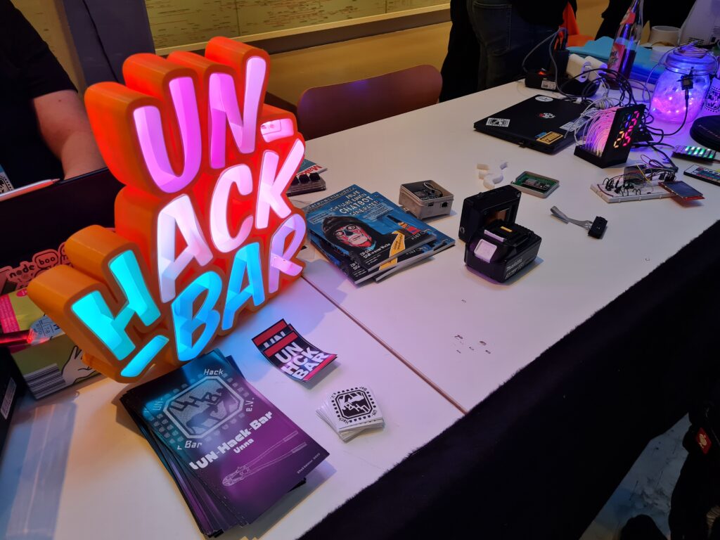 Ein Tisch mit einer Art Neon-Schriftzug "UN-Hack-Bar", Flyer, Aufkleber, Datenschleuder, ein Makita-Akku, Platinen in einer Aufputzdose, Eine bunte 7-Segment-Uhr, ein Laptop, ein Steckbrett mit nicht näher erkennbaren Projekt, ein Einkochglass mit LEDs und ein geschlossener Laptop.