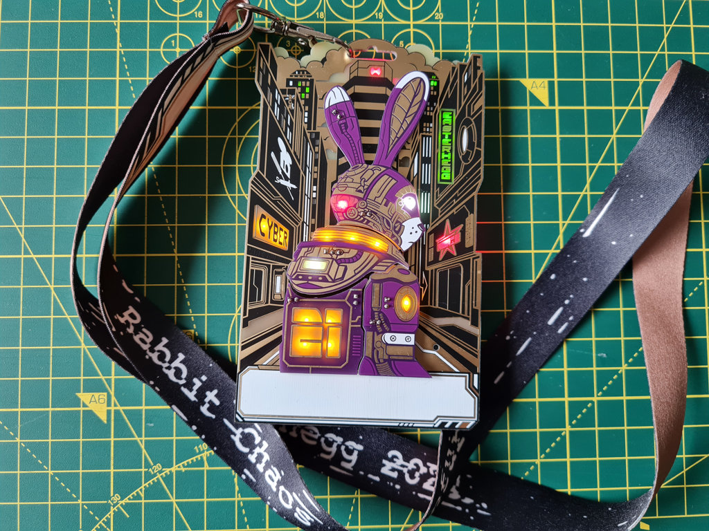 Ein PCB-Badge mit einem "Cyber-Hasen" auf der Vorderseite. Der Hase steht in einer Art futuristschen Großstadt. Von hinten leuchten div. farbige LEDs durch die Platine und beleuchten so div. Elemente (Auge des Hasen, ein "Neuralknoten" am Hinterkopf, Symbole auf der Jacke des Hasen, Leuchtreklamen und ein Freifunk-Logo auf dem Dach von einem Hochhaus)