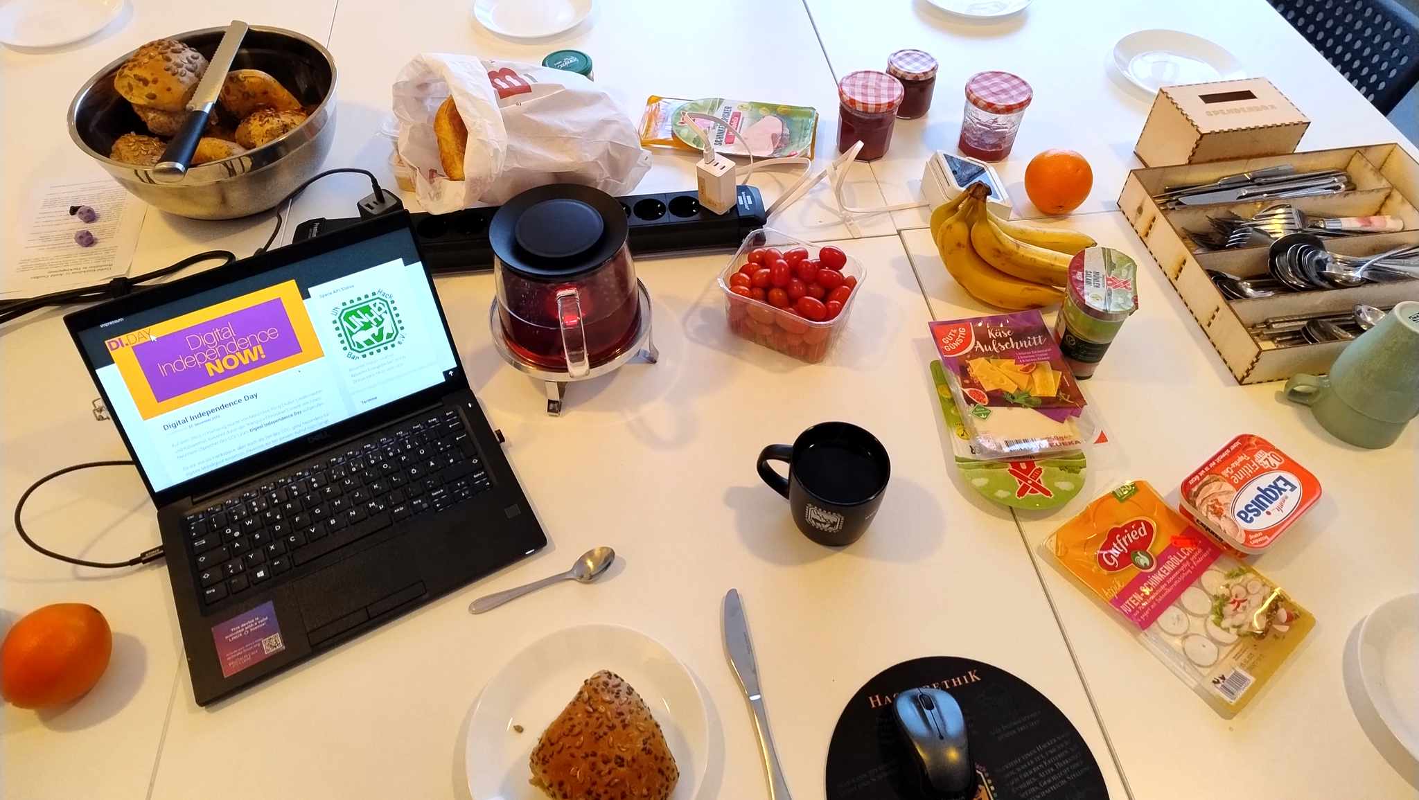 Ein gedeckter Frühstückstisch mit Brötchen, div. Belagen, Tassen, Teller, Besteckkasten usw. Außerdem ein Laptop der die UNHB-Webseite mit einem DI-Day Blogpost anzeigt.