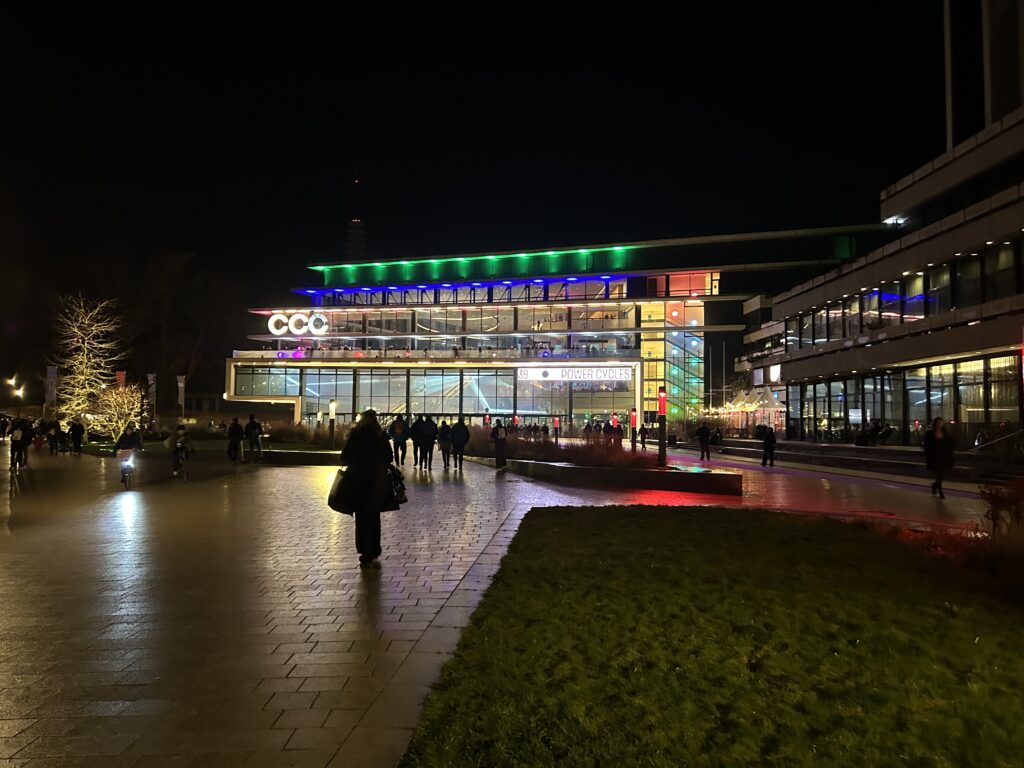 Der Platz vor dem Eingang des CCH zu später, dunkler Stunde. Man sieht die Fassade des Gebäudes mit vielen bunten Lichtern und Leuten die ins Gebäude gehen oder es verlassen. Der Schriftzug "CCH" auf dem Gebäude wurde verändert so das dort nun "CCC" steht.