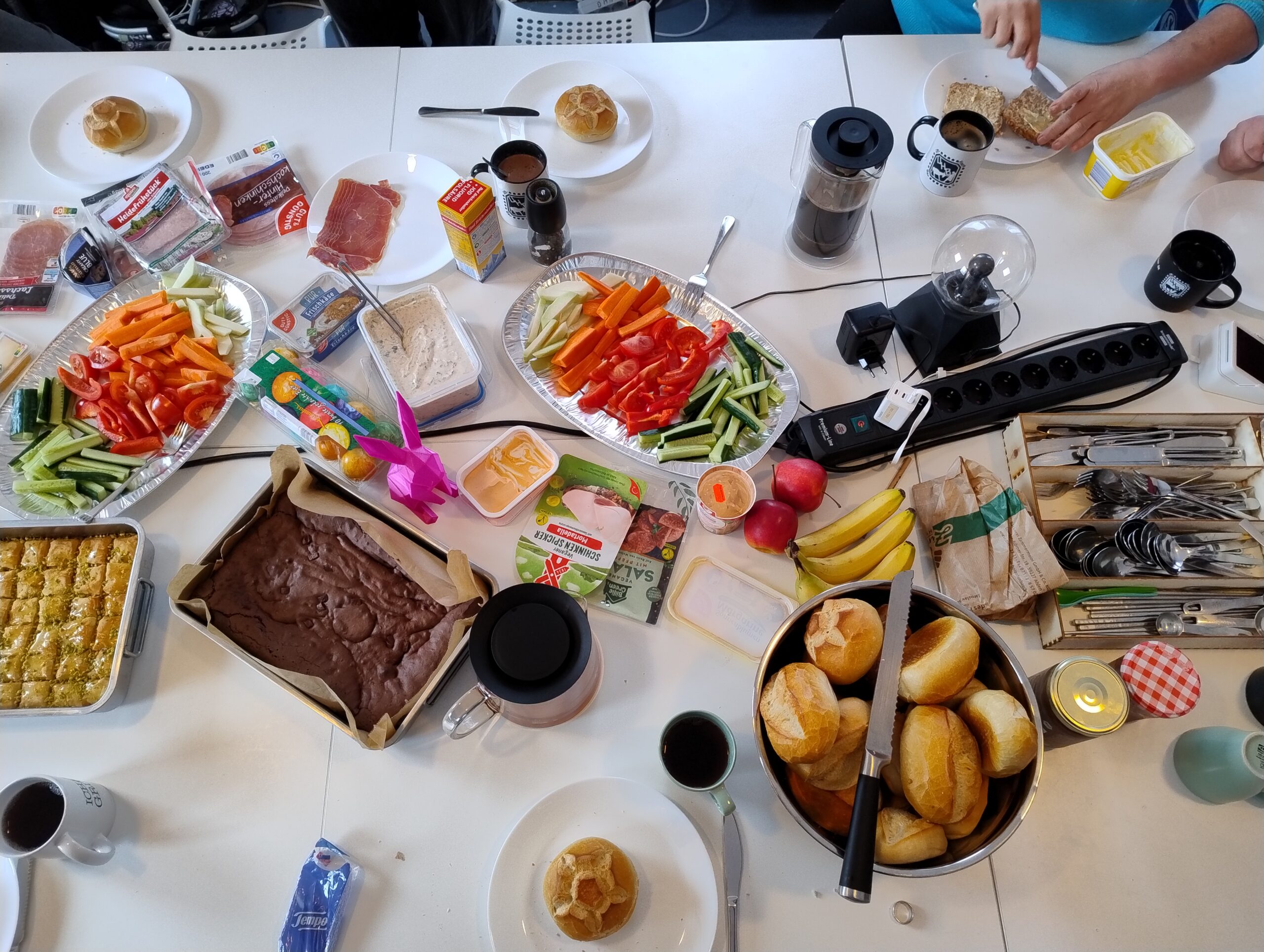 Ein reich gedeckter Frühstückstisch. Man sieht eine Schüssel voll Brötchen, eine Rohkostplatte, Obst, Aufstrich, Dips, Marmeladen, Kuchen, Tee, und Kaffee